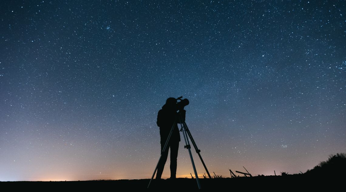 A person stands on a hill, gazing through a telescope, surrounded by a scenic landscape under a clear sky.